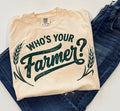 A folded cream-colored t-shirt with a dark green graphic reading 'WHO'S YOUR Farmer?' with wheat illustrations, styled on top of blue jeans against a cream background.