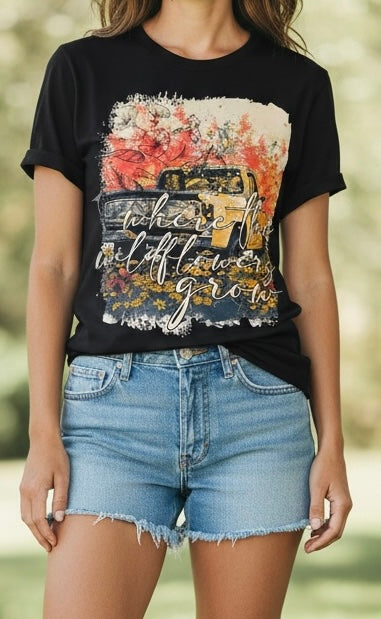 A woman wearing a black graphic t-shirt with a vintage truck and floral design, paired with light-wash denim shorts, set against a blurred outdoor background.