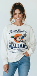 A woman wearing a cream-colored crewneck sweatshirt with a vintage-style mallard duck graphic and text reading 'Rusty Feather Outfitters' and 'Mallard Brand Co. American Hunters,' paired with blue jeans, against a light gray studio background.