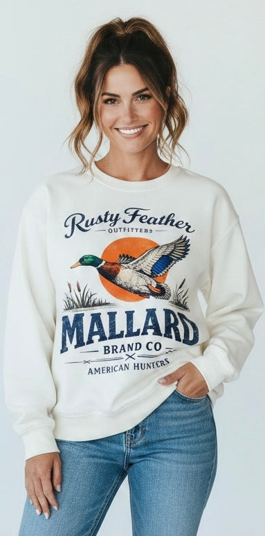 A woman wearing a cream-colored crewneck sweatshirt with a vintage-style mallard duck graphic and text reading 'Rusty Feather Outfitters' and 'Mallard Brand Co. American Hunters,' paired with blue jeans, against a light gray studio background.