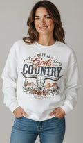 A woman wearing a white crewneck sweatshirt with a graphic print that reads 'THIS IS God's COUNTRY' with a longhorn skull and desert landscape illustration, paired with blue jeans against a neutral gray background.