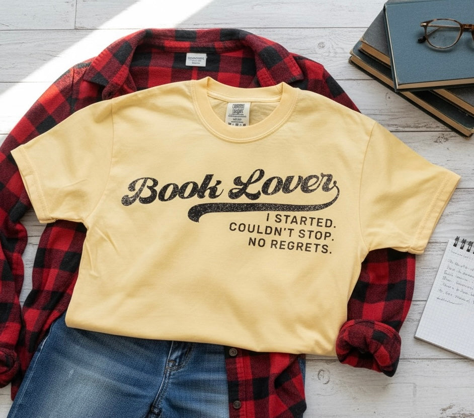 A pale yellow graphic T-shirt laid flat on top of a red-and-black plaid flannel shirt, with blue jeans, books, and glasses around it on a white wood surface. The shirt reads 'Book Lover' in large black script with the phrase 'I STARTED.