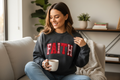 A woman sitting on a beige couch in a cozy living room setting, wearing a dark gray crewneck sweatshirt with the word 'FAITH' in red letters outlined with white dots. She is holding a white mug.