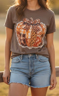 A woman wearing a brown short-sleeve t-shirt with a large autumn-themed pumpkin graphic on the front, paired with light-wash denim shorts.