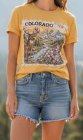 A woman wearing a mustard yellow graphic t-shirt with a 'COLORADO' deer and mountain print, paired with frayed denim shorts.