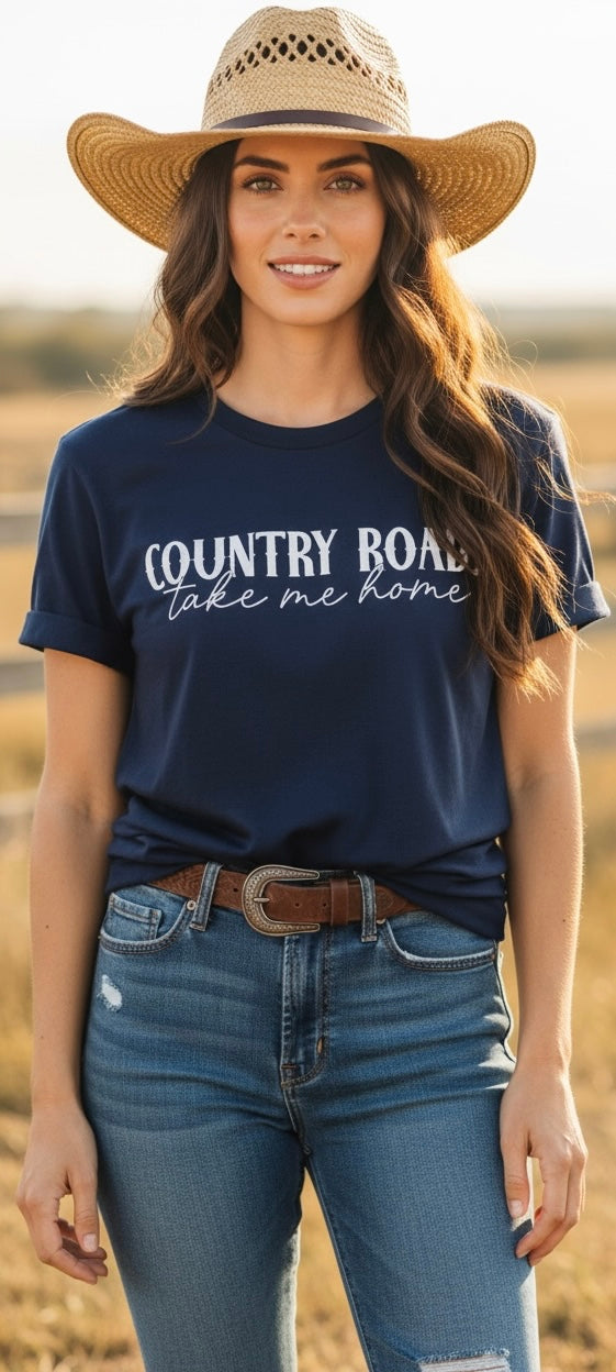 A woman wearing a navy blue graphic t-shirt with 'COUNTRY ROAD take me home' printed in white script, paired with a straw cowboy hat and blue jeans, standing in a dry field.