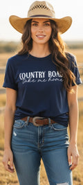 A woman wearing a navy blue graphic t-shirt with 'COUNTRY ROAD take me home' printed in white script, paired with a straw cowboy hat and blue jeans, standing in a dry field.