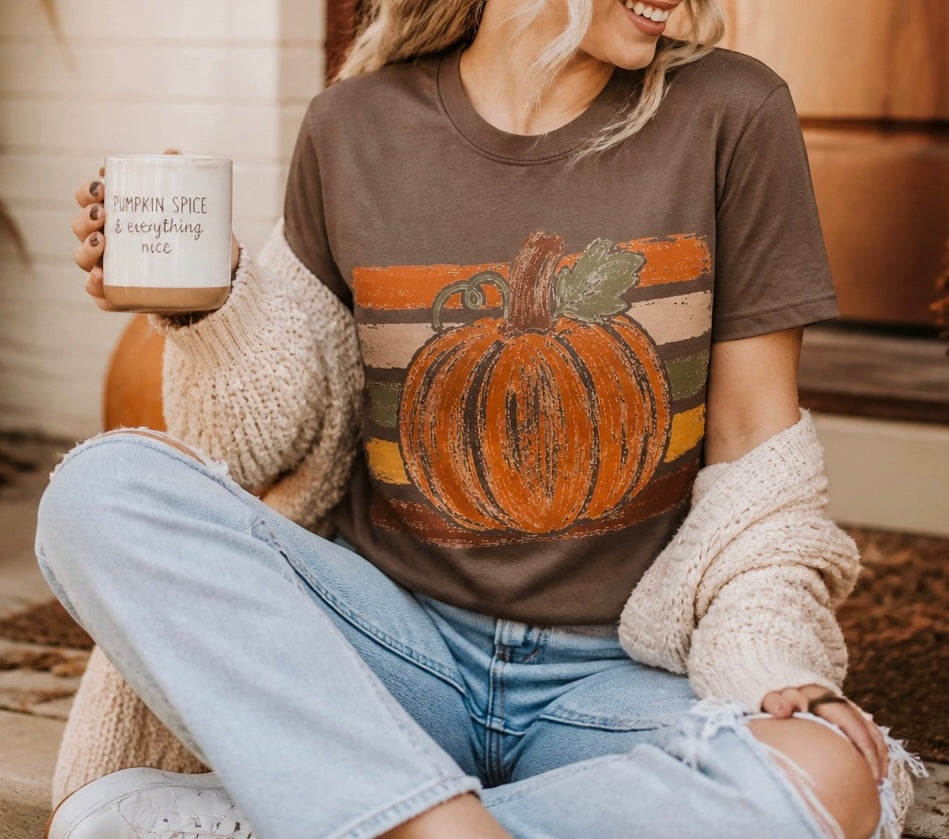 A woman wearing a brown graphic t-shirt with a large orange pumpkin design, styled with a chunky cream knit sweater and light-wash jeans, holding a mug that reads 'PUMPKIN SPICE & everything nice'.