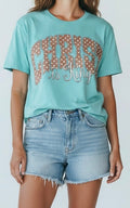 Person wearing a teal t-shirt with  'Christ is King' text and denim shorts on a plain background
