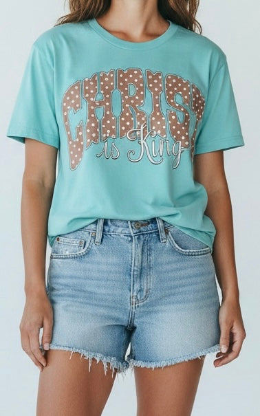 Person wearing a teal t-shirt with  'Christ is King' text and denim shorts on a plain background