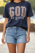 A woman wearing a navy blue graphic t-shirt with 'GOD IS good' printed on the front, where 'GOD' features a leopard print pattern. She is also wearing light wash denim shorts.