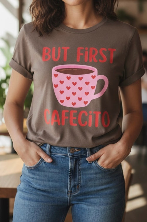 A woman wearing a brown graphic t-shirt with the text 'BUT FIRST CAFECITO' and a pink coffee mug with red hearts, paired with blue jeans.