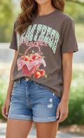 A woman wearing a brown graphic t-shirt with a 'Sweetest Peaches' print and distressed denim shorts, photographed outdoors with a blurred green background.