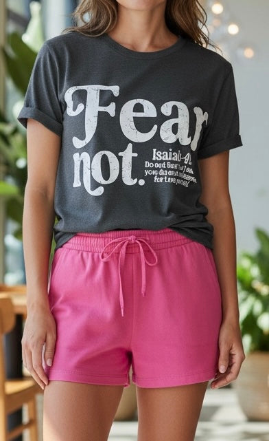 A woman wearing a dark charcoal gray graphic t-shirt with the text 'Fear not. Isaiah 40:29' in white, paired with bright pink drawstring shorts.