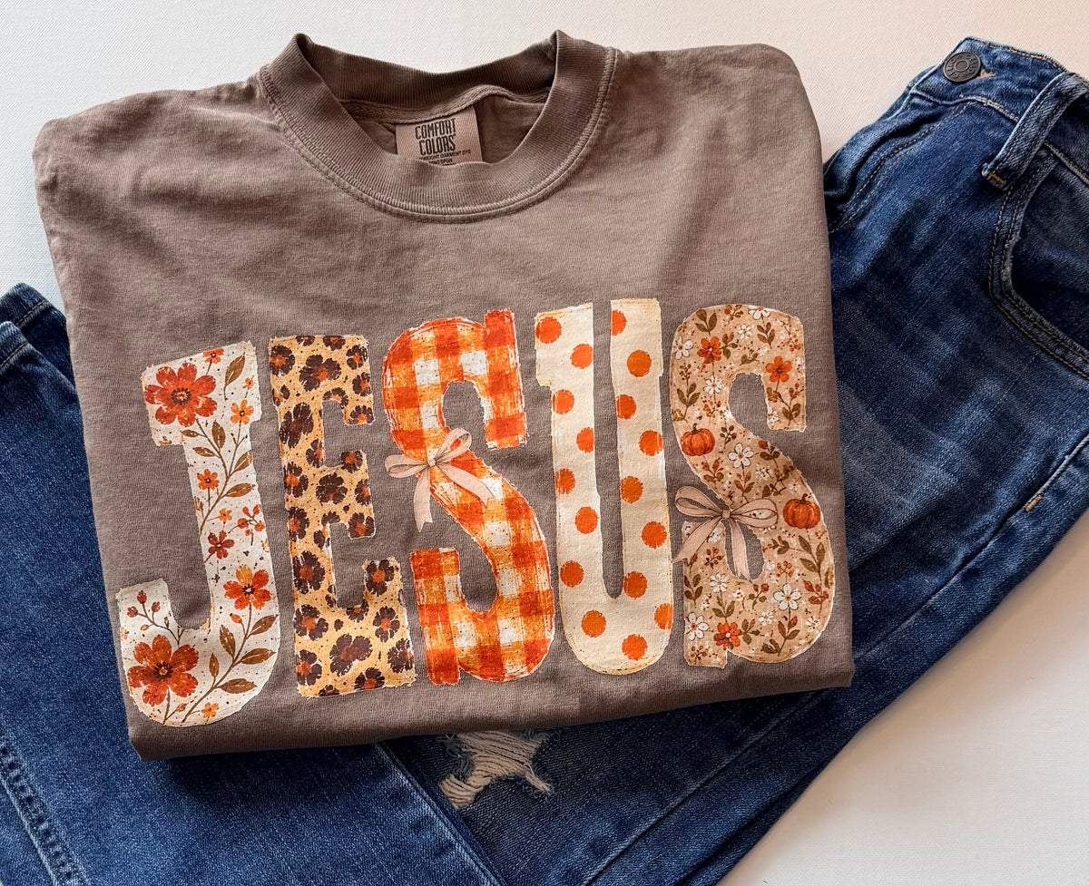 A folded taupe gray t-shirt with the word 'JESUS' printed across the front in decorative autumn-themed patterns, styled on top of blue jeans.