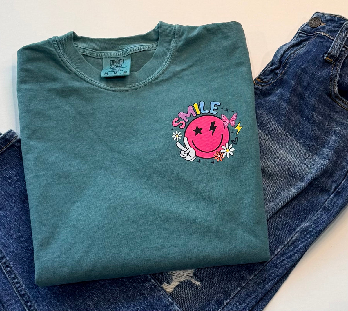 A folded sage green t-shirt with a colorful 'SMILE' graphic on the chest, styled on top of blue jeans against a white background.
