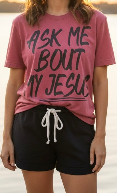 A woman wearing a dusty rose graphic t-shirt with the text 'ASK ME BOUT MY JESUS' in black lettering, paired with black drawstring shorts, set against a soft-focus outdoor background.