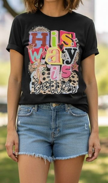 A woman wearing a black graphic t-shirt with the text 'His Way Is Better' in a colorful collage design, paired with denim shorts, set against a blurred outdoor background.