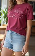Person wearing a maroon t-shirt with a mountain design and text, paired with denim shorts indoors.