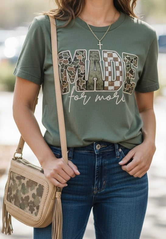 A woman wearing an olive green graphic t-shirt with 'MADE for more' in camouflage and checkered lettering, paired with blue jeans and a beige camouflage crossbody bag.
