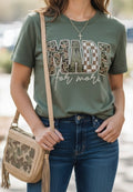 A woman wearing an olive green graphic t-shirt with 'MADE for more' in camouflage and checkered lettering, paired with blue jeans and a beige camouflage crossbody bag.