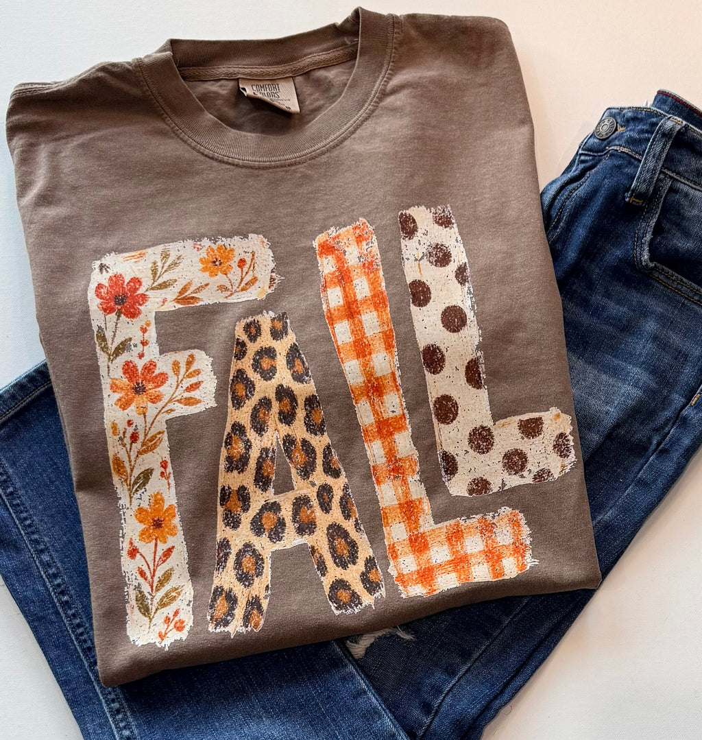 A folded taupe-brown t-shirt with a distressed 'FALL' graphic featuring leopard print, orange gingham, and floral patterns, styled on top of blue jeans.