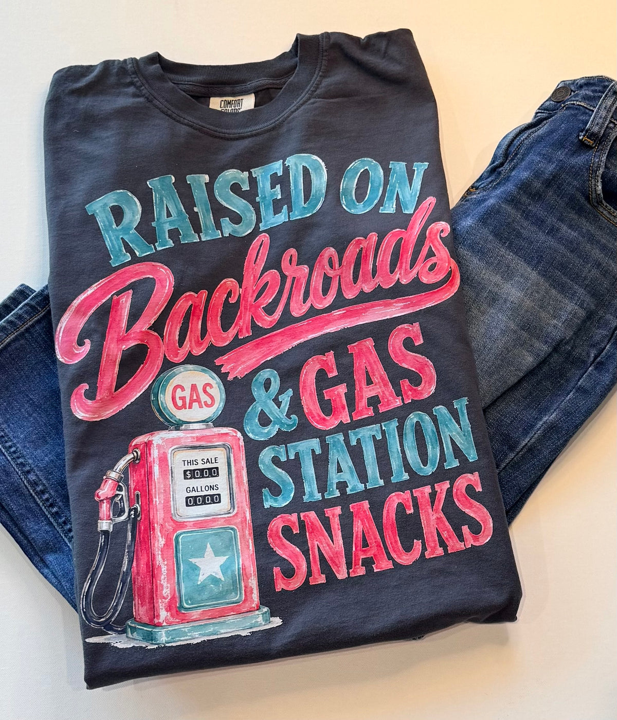 Black t-shirt with colorful graphic and text 'Raised on Backroads & Gas Station Snacks' on a white background.