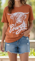 Person wearing a brown t-shirt with a cow graphic and 'Cowgirl Say' text, paired with denim shorts.