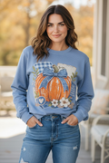 A woman wearing a dusty blue crewneck sweatshirt with a large autumn-themed pumpkin graphic on the front, paired with blue jeans, standing outdoors on a porch.
