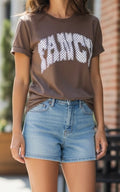 A woman wearing a brown short-sleeve graphic t-shirt with 'FANCY' printed in white polka-dot letters, paired with light-wash denim shorts, shown in an outdoor setting.