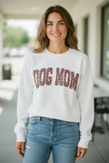 A woman wearing a white crewneck sweatshirt with 'DOG MOM' printed across the chest in leopard-print letters, paired with light-wash blue jeans, standing in a brightly lit, modern indoor hallway.