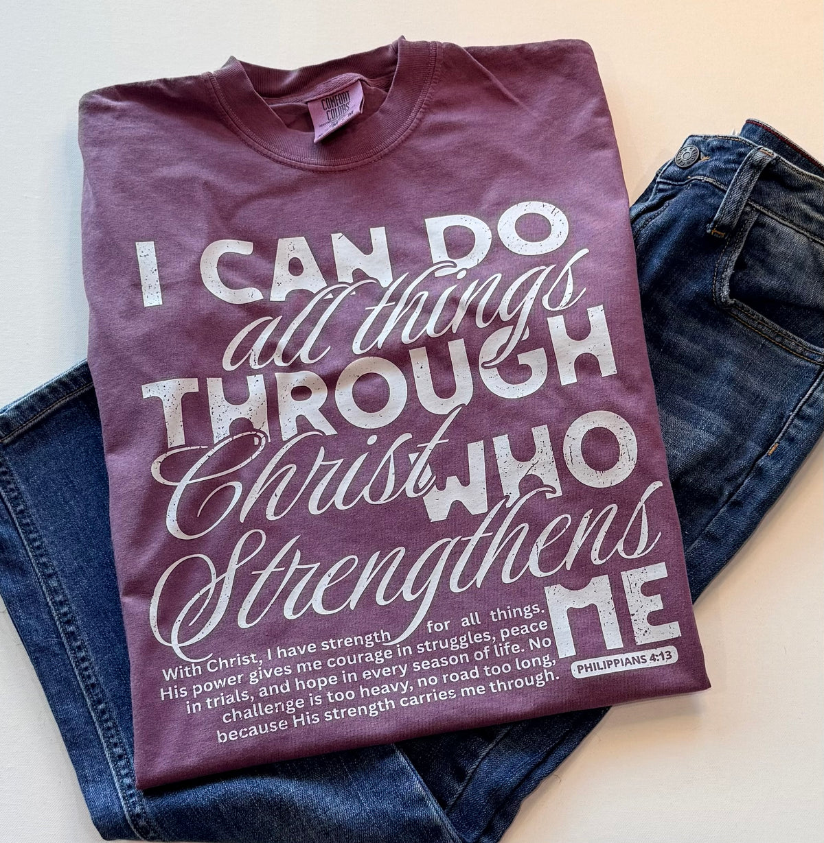 A folded maroon t-shirt with a white printed Bible verse about strength through Christ, styled on top of a pair of blue jeans against a light beige background.