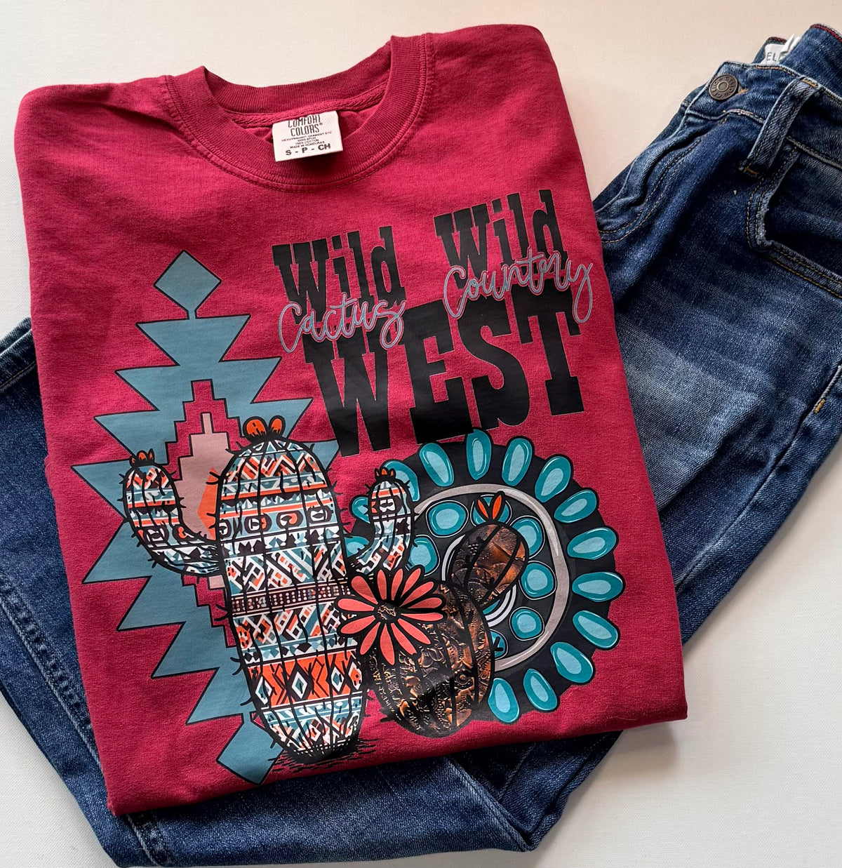A folded maroon graphic T-shirt with a southwestern design featuring a patterned cactus, turquoise geometric shapes, and the words 'Wild West' and 'Cactus Country,' styled on top of blue jeans against a light background.
